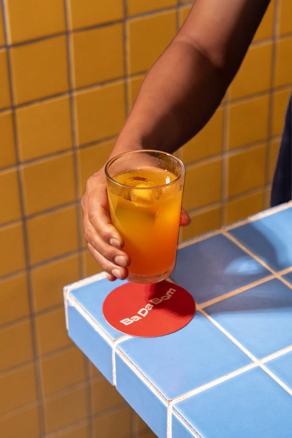 A hand holding an orange drink above a red coaster on blue tiles