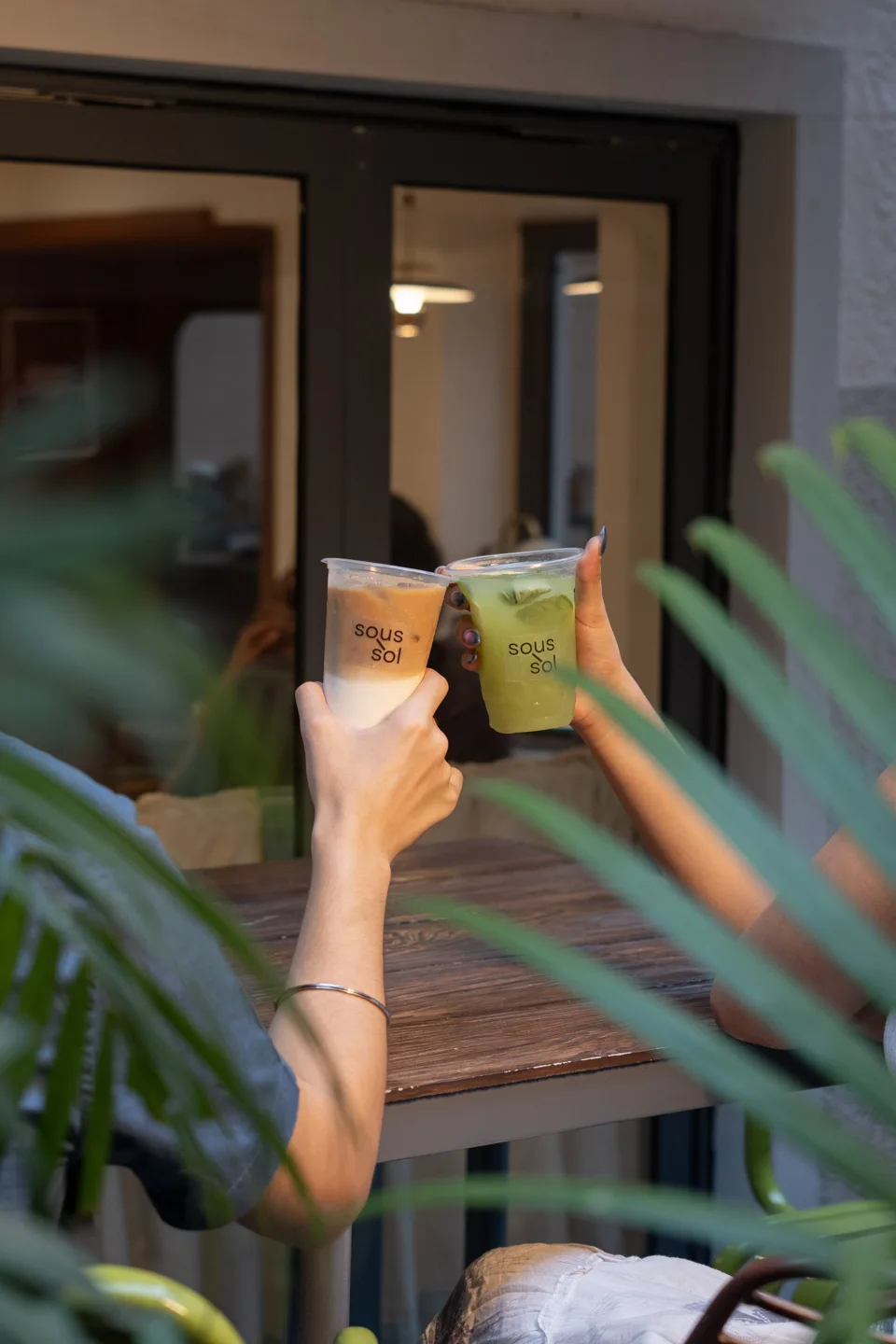 A hand holding a drink beside green café plants
